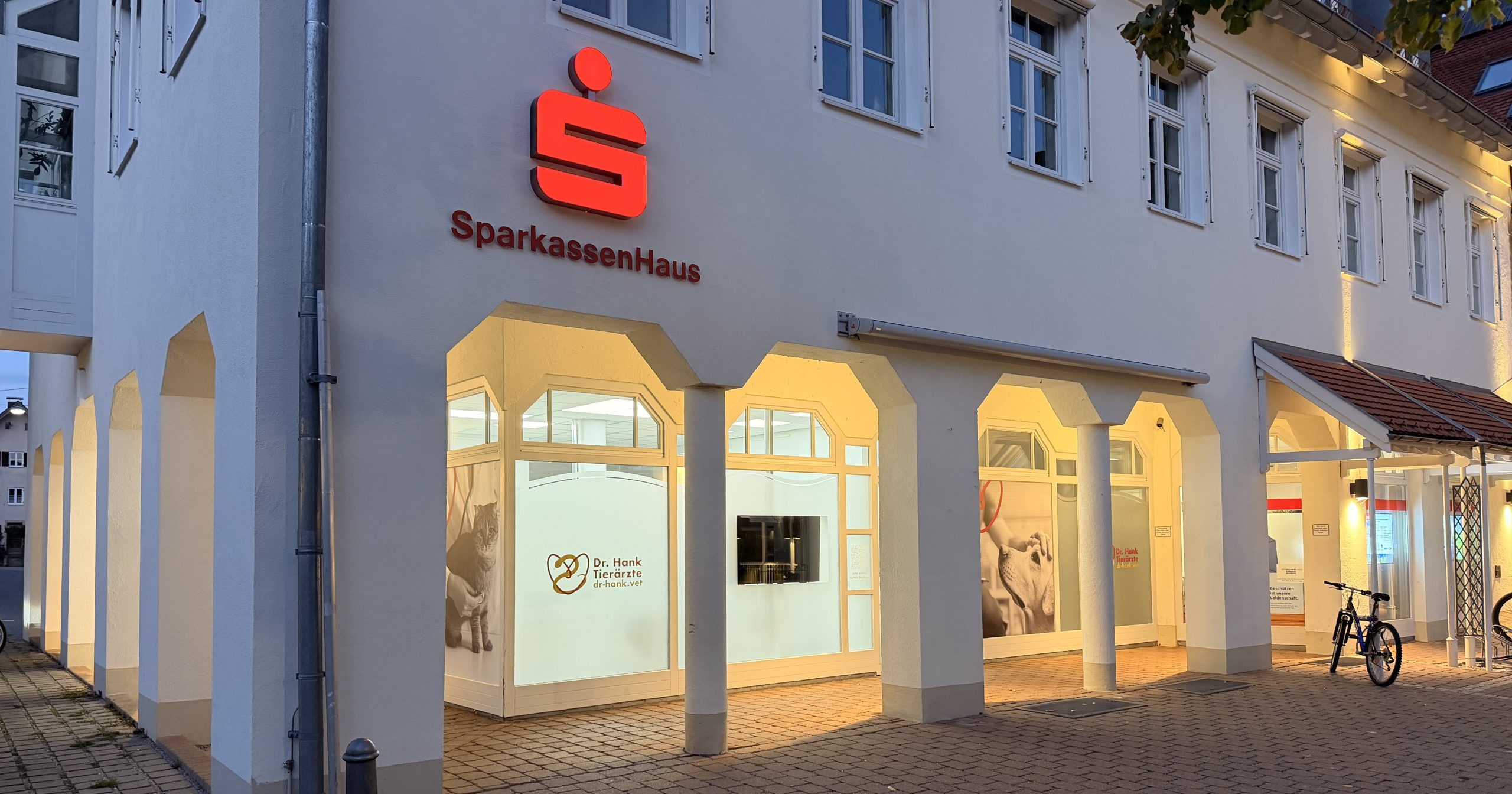 Die Kleintierpraxis Peiting am Abend: beleuchtete Schaufenster unter den Arkaden des SparkassenHauses am Hauptplatz
